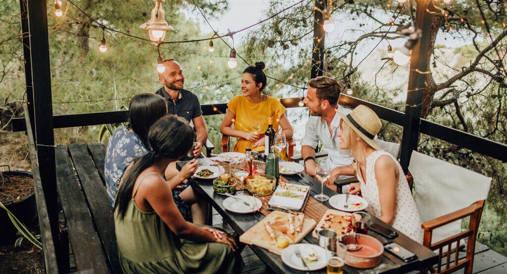 Group of friends laughing around a long wooden table with food and wine on a pergola terrace under string lights, surrounded by pine trees and sea views