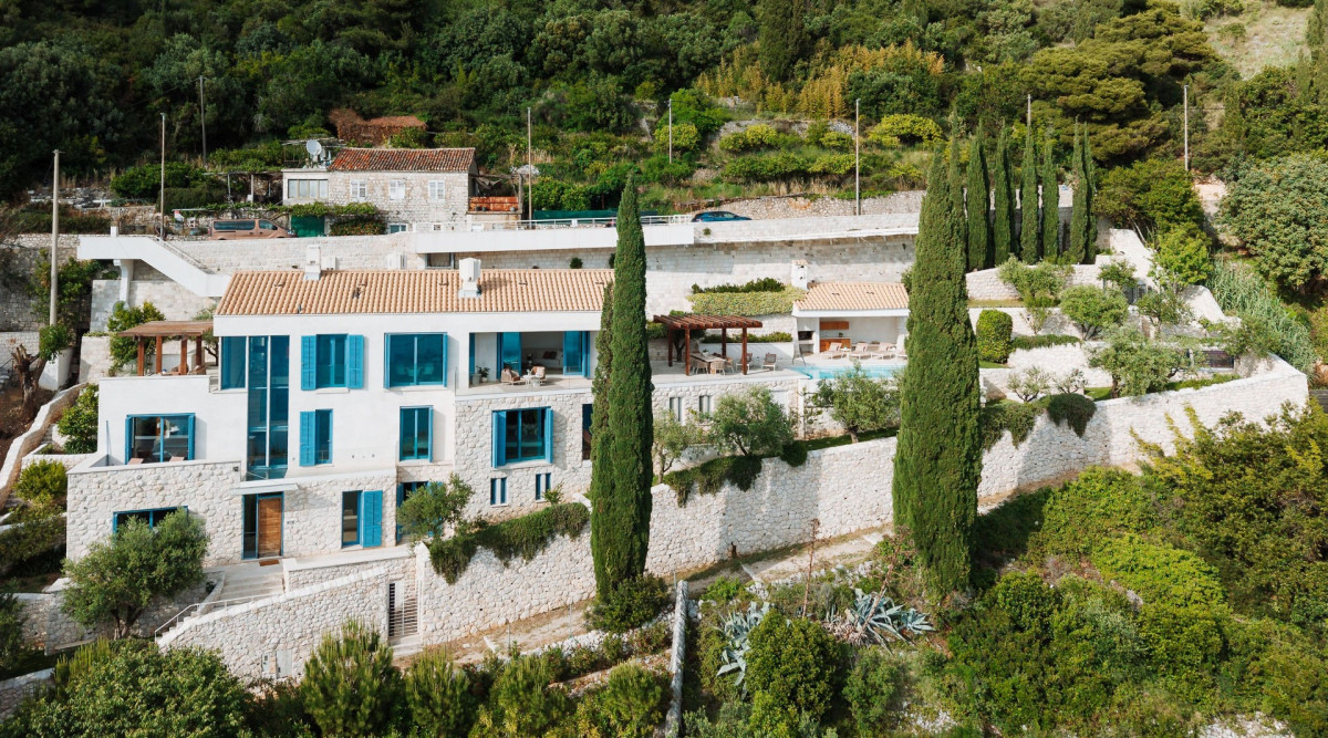Außenansicht der Villa Blazia in Dubrovnik mit blauen Rollläden, einer Terrassenanlage und Zypressen auf dem Hügel