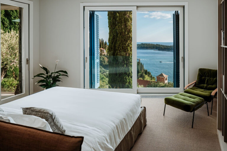 Schlafzimmer in der Villa Blazia mit einem Doppelbett, einer grünen Sesselstühle und einem Balkontür mit Blick auf das Meer
