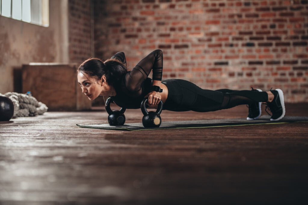 Woman in black sportswear doing a push-up on kettlebells on a mat in a loft-style gym with a brick wall